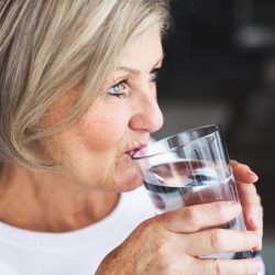 Alte Frau beugt Ihren Kopfschmerzen vor und trinkt viel Flüssilgkeit in FOrm von Wasser